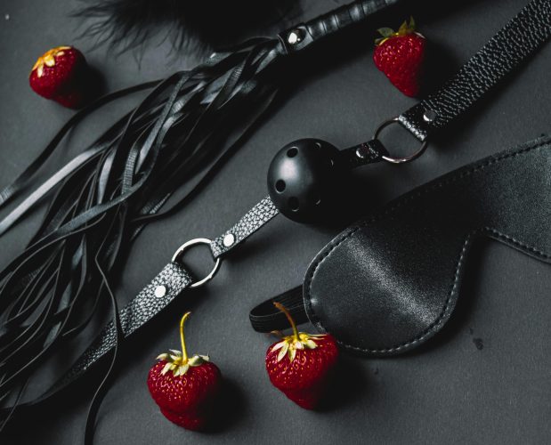 Flat lay of black leather BDSM items with fresh strawberries on dark background.